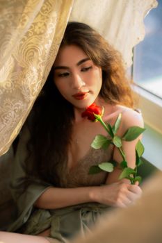 Romantic portrait of a woman holding a red rose, bathed in soft natural light by a window.