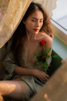 A peaceful moment as a woman holds a red rose by the window, bathed in soft sunlight.