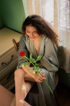 Serene woman in a silk robe holding a red rose, sitting by a sunlit window, reflecting calmness.