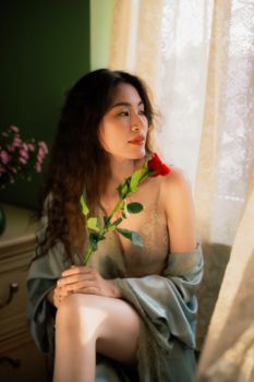 A serene portrait of a young woman in soft light holding a red rose by the window.