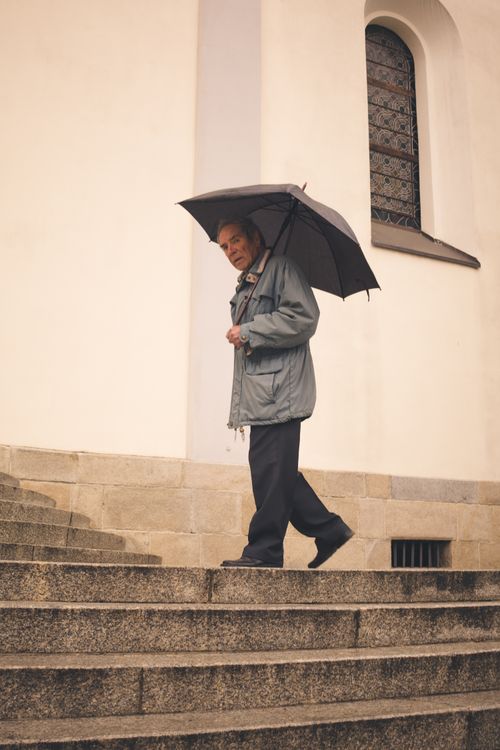Free Elderly man in coat and umbrella walking down steps near building exterior. Stock Photo