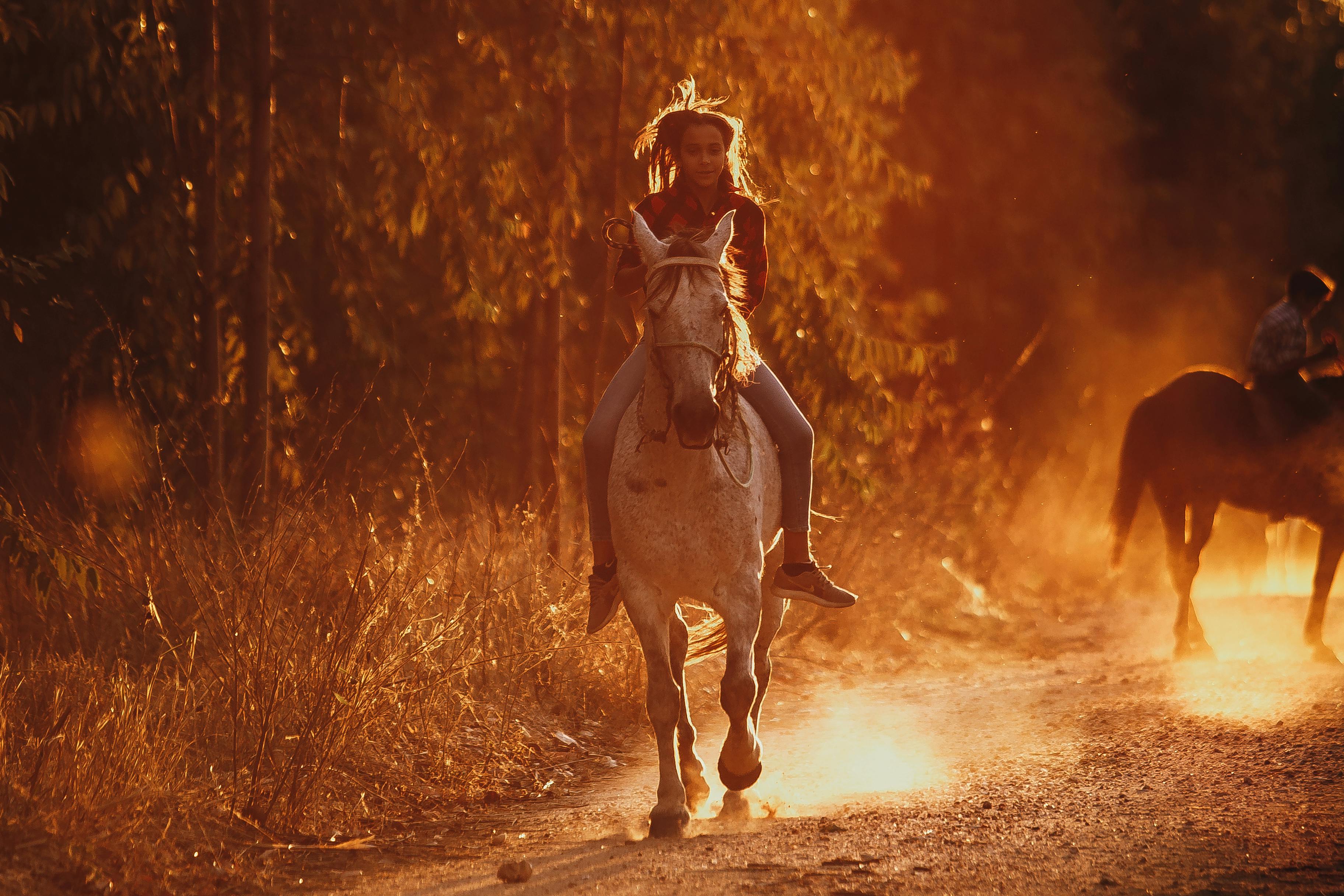 A woman rides a horse on a dusty trail during sunset, creating a warm, atmospheric scene.