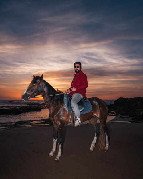 Free A man in a red shirt rides a horse by the ocean at sunset, capturing a serene coastal scene. Stock Photo