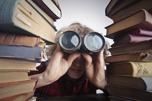 Anonymous person with binoculars looking through stacked books