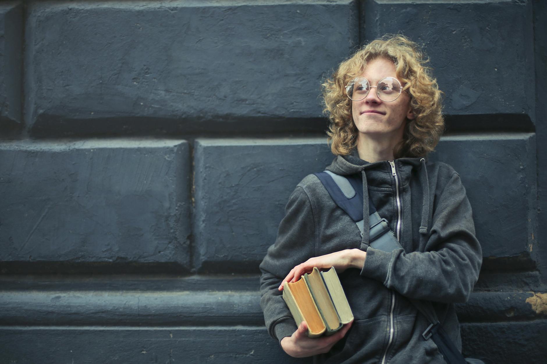 A smiling young man with glasses and messy hair holds books, leaning against a textured wall.