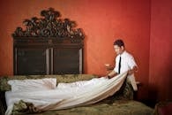 Side view of young ethnic male making bed while cleaning stylish hotel room with vintage furniture and red walls