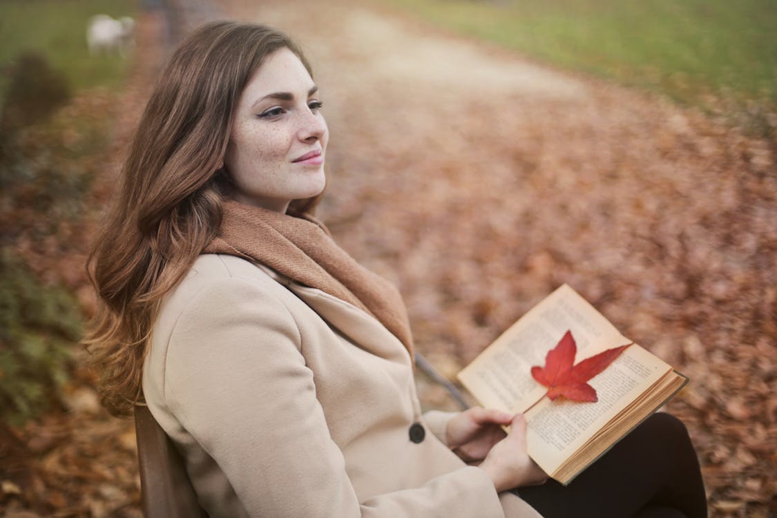 Side view of peaceful young female in warm coat and scarf sitting on bench with open book in hands and looking away pensively while resting in autumn park