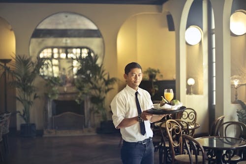 Free Confident young ethnic waiter in elegant clothes holding tray with food and drinks and looking at camera while serving tables in stylish restaurant Stock Photo