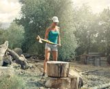 Young female in casual clothes standing in front of large log with axe in hands while chopping wood in backyard of country house in sunny summer day