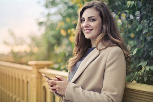 Selective Focus Photo of Smiling Woman in Brown Coat Holding Smartphone While Leaning on stone Railing