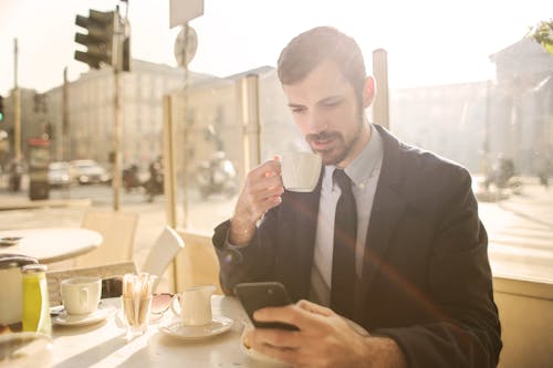 穿著黑西裝的男人在一家咖啡館喝咖啡時使用他的電話的照片