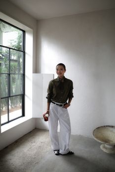 Young woman in casual attire standing by a large window with natural light, holding a drink.