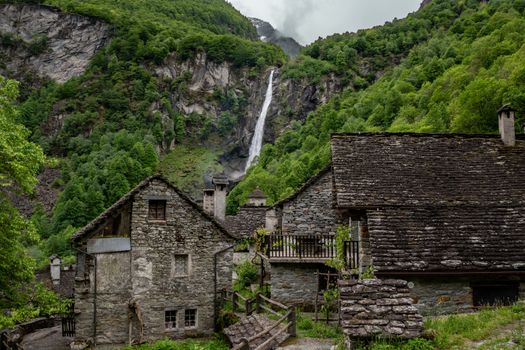 Idyllic view of stone houses in Cevio, Ticino, with lush greenery and a dramatic alpine waterfall.