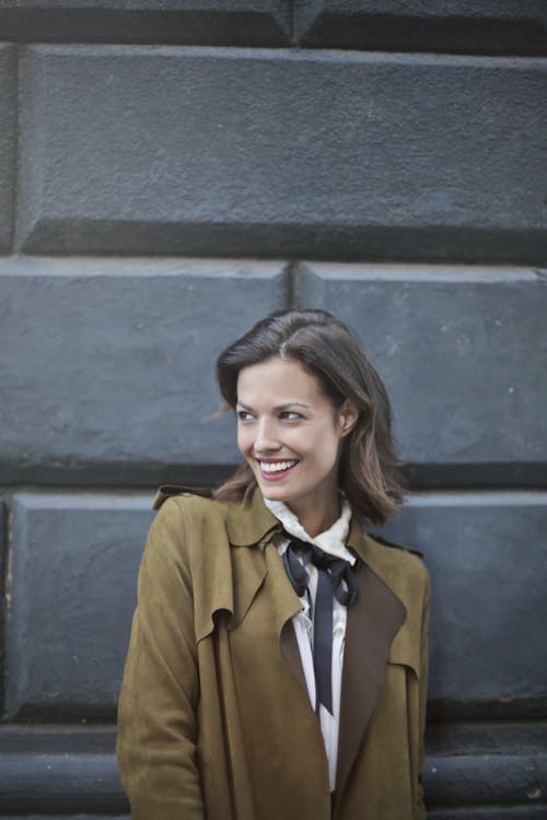 Женщина в коричневом пальто стоит возле серой бетонной стены