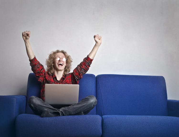 Photo Of Excited Person With Hands Up Sitting On A Blue Sofa While Using A Laptop