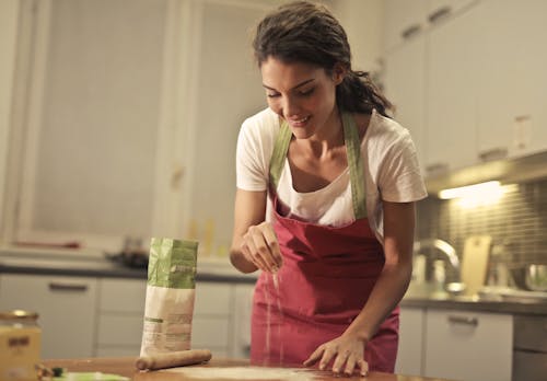 Imagen de una joven positiva en un delantal espolvoreando harina sobre la mesa mientras prepara comida en una cocina moderna y luminosa