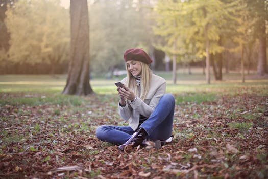 一名妇女坐在公园里，周围满是秋叶和树木，正用着她的智能手机
