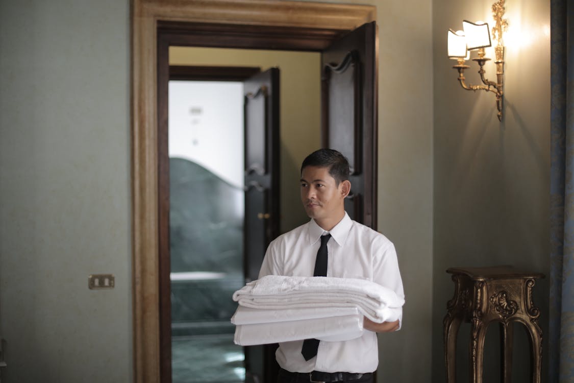Young male housekeeper carrying stack of white bed sheets while entering bedroom in hotel