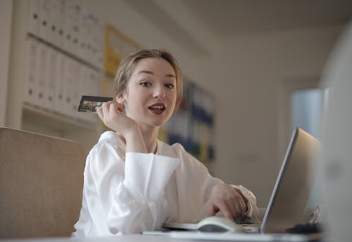 Low angle side view of positive young female entrepreneur in formal wear using laptop and performing online payment with credit card while sitting at table at workplace