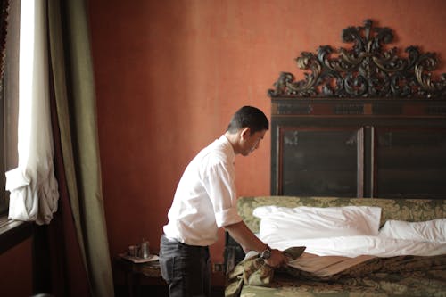 Free Side view of young male housekeeper making bed while arranging stylish hotel bedroom with vintage interior Stock Photo