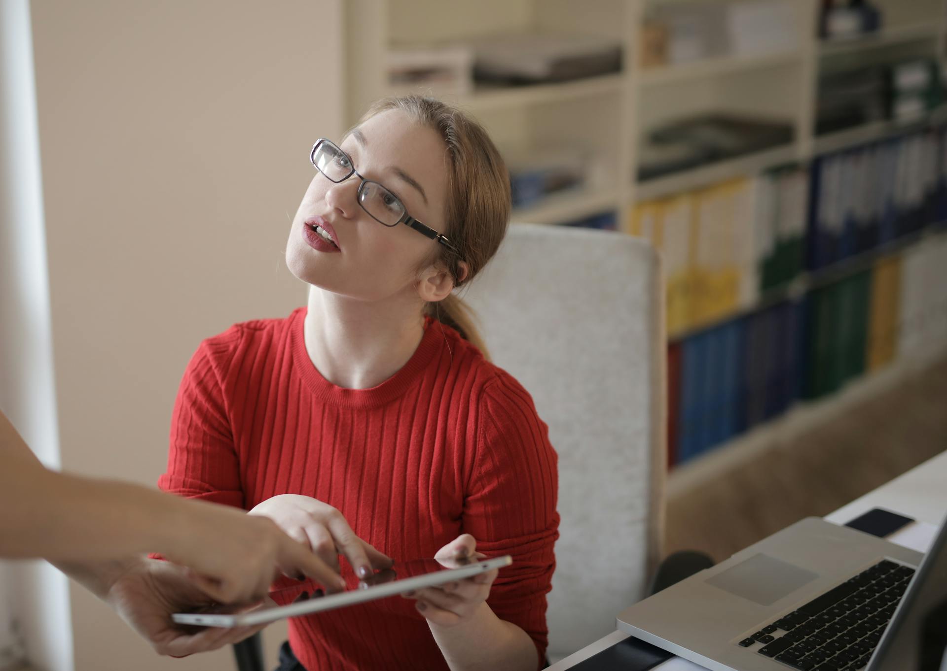 Professional woman in red sweater discussing data on a tablet in office setting, working together.