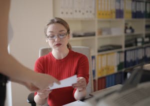 Serious young female employee in casual clothes and eyeglasses reading attentively document while working in office with colleague