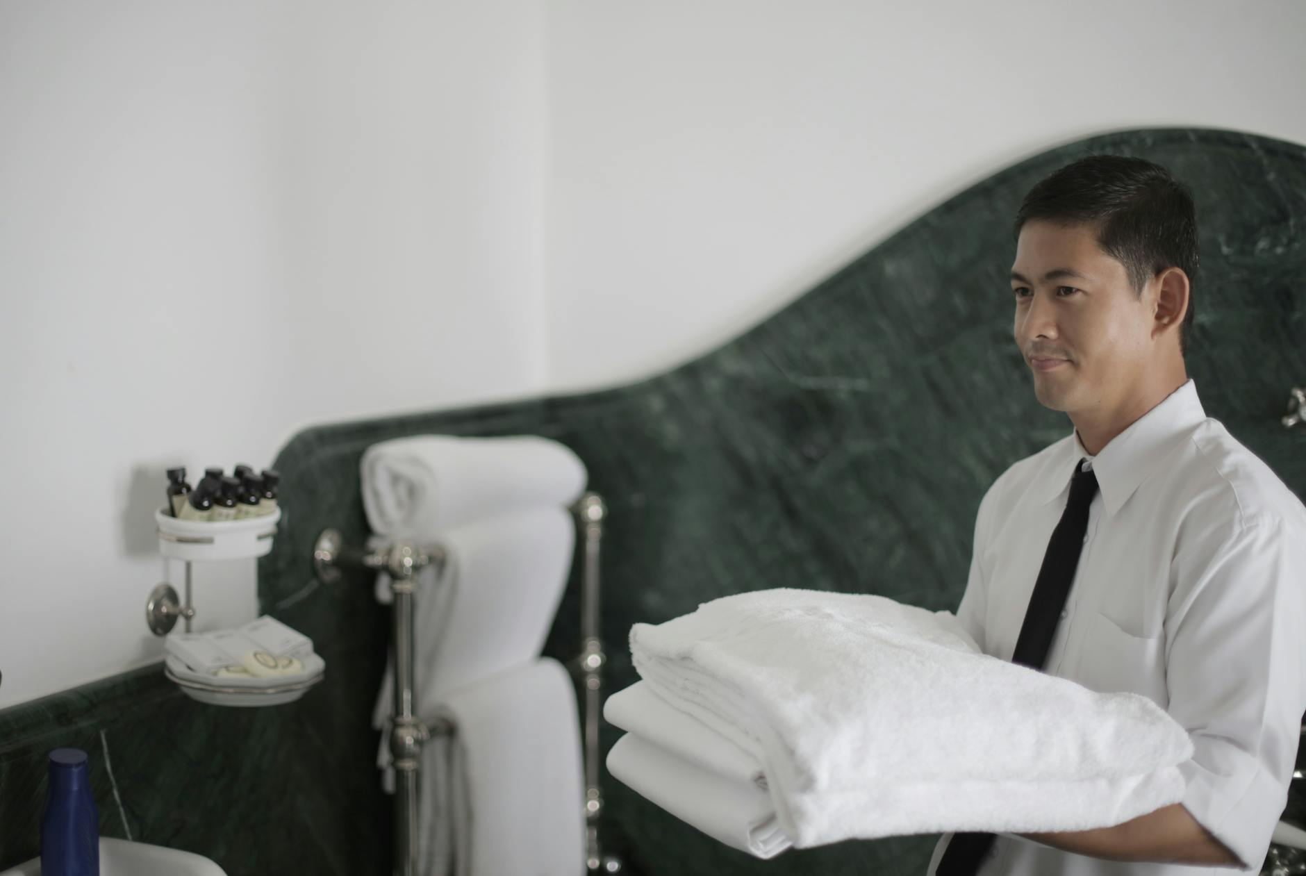 Side view of smiling ethnic male hotel employee holding stack of clean white towels while cleaning bathroom