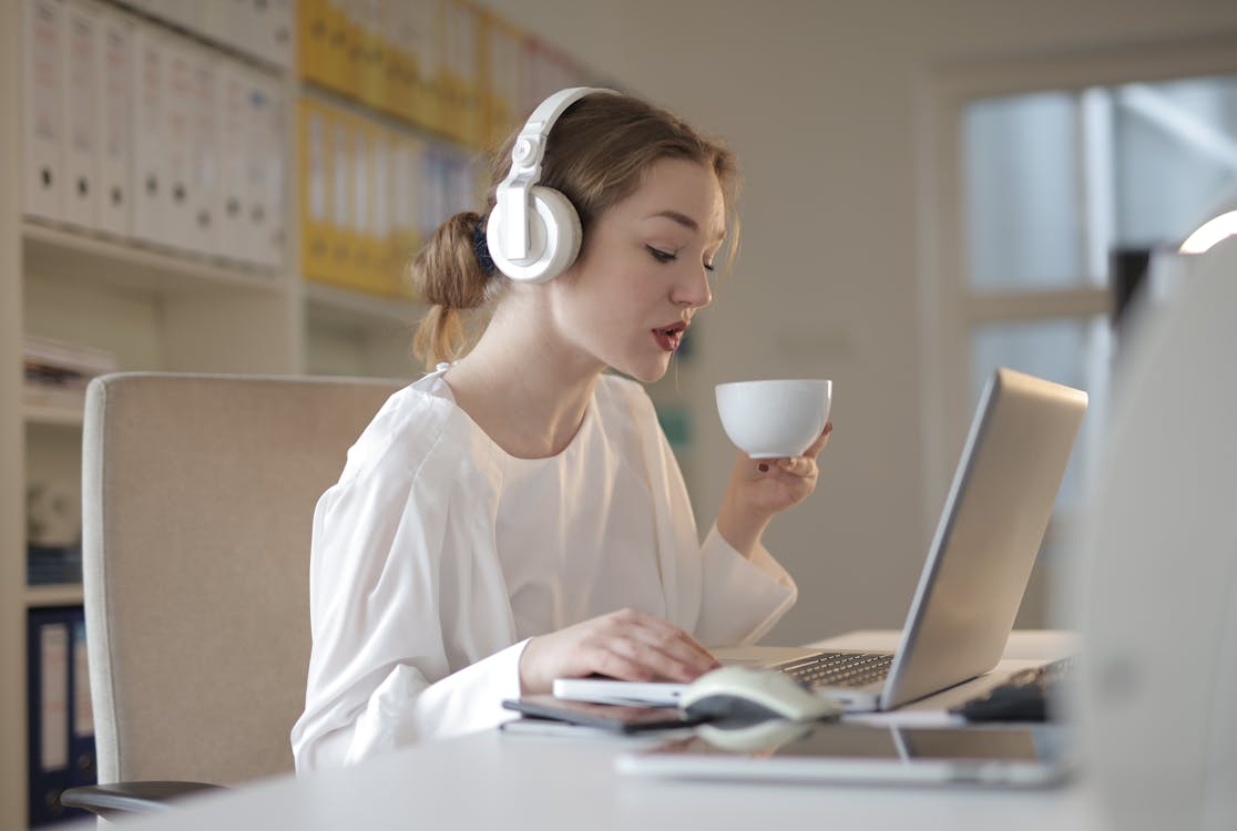 Selective Focus Photo of Woman in Headphone Listening to Music While Drinking Coffee and Using Her Laptop