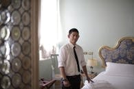 Young ethnic male attendant standing next to bed with stacked linen in stylish hotel room and looking at camera