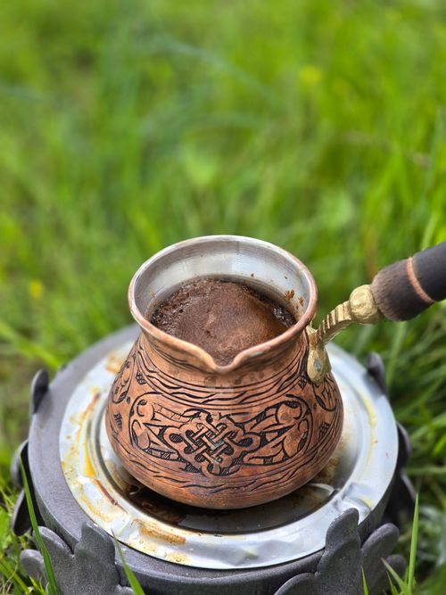 Free A decorative copper pot with coffee simmering on an outdoor stove, set in a grassy field. Stock Photo