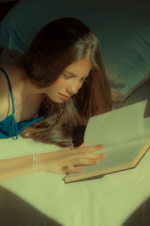 Free A young woman with long hair enjoying a book while lying in bed under soft sunlight. Stock Photo