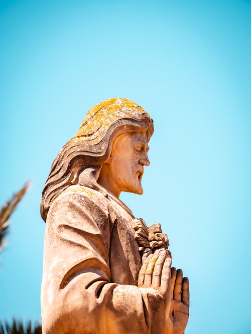 Free A stone statue of Jesus in prayer, shot under a clear blue sky creating a serene atmosphere. Stock Photo