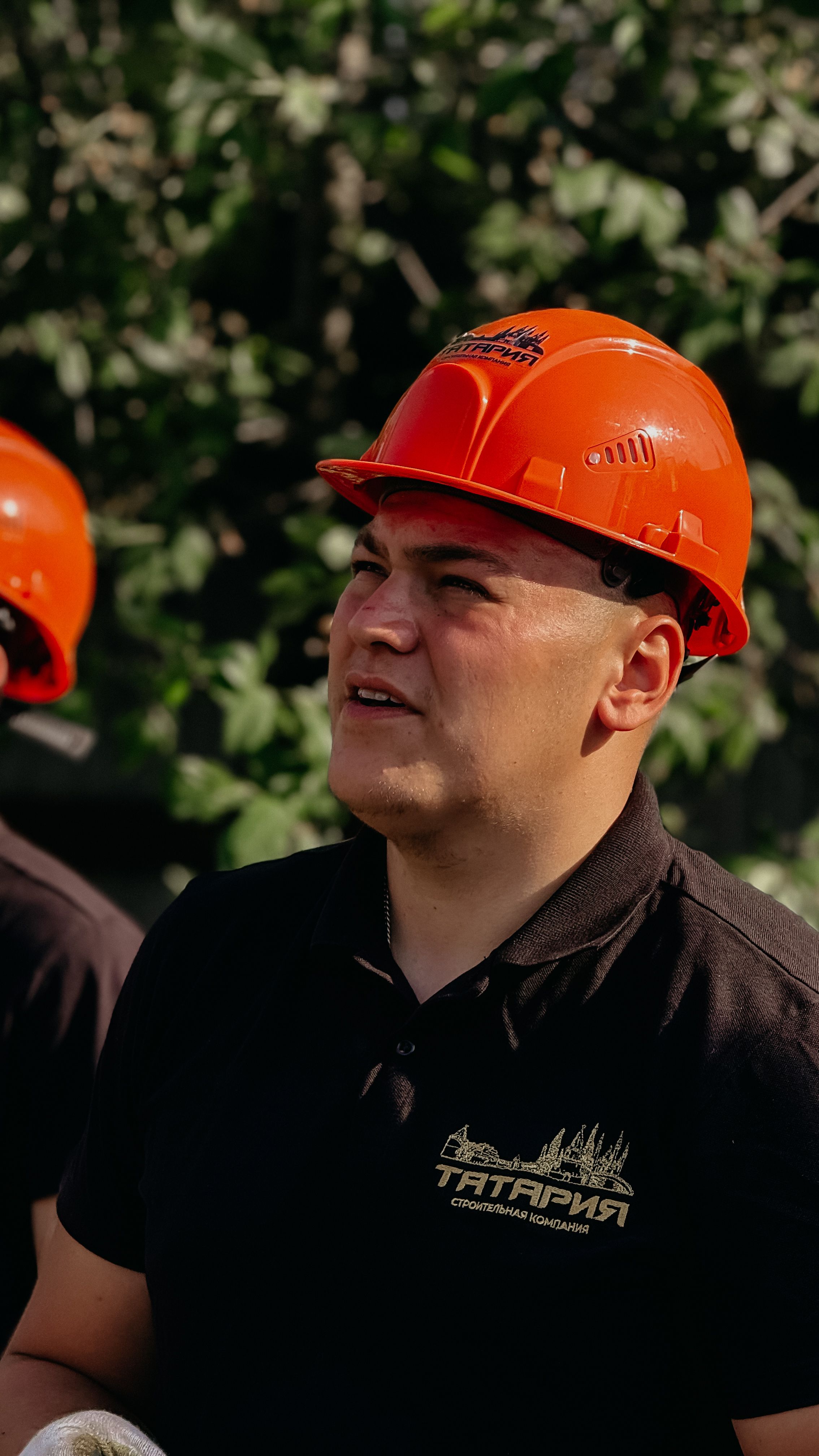 A construction worker wearing a red hard hat and black shirt, standing outdoors amidst trees.