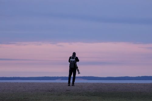 Free Silhouetted photographer takes a picture by the beach at twilight under pastel skies. Stock Photo