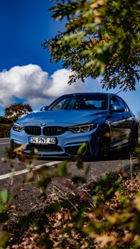 A sleek blue sedan drives on a scenic road surrounded by lush greenery under a vibrant blue sky.