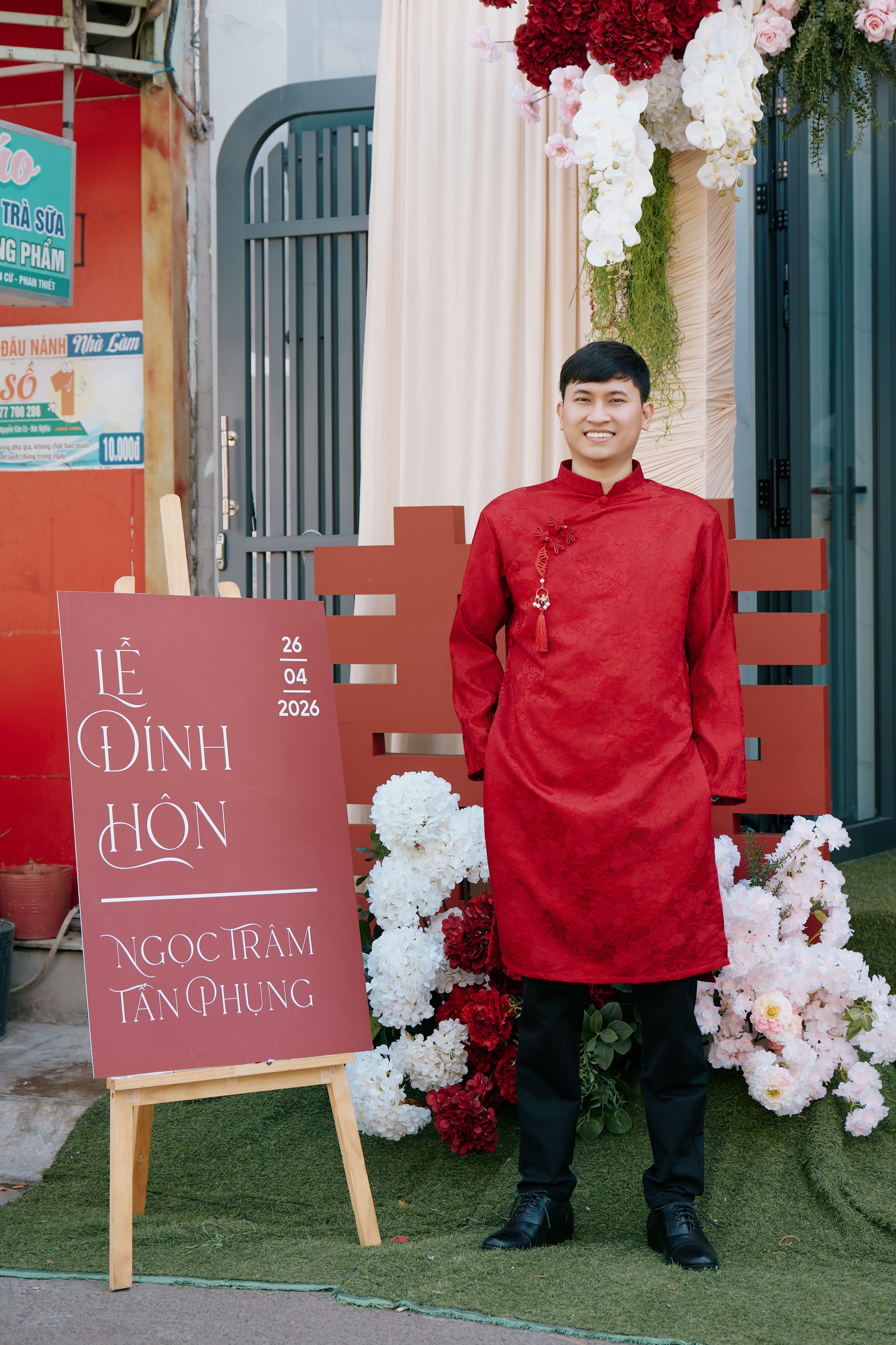 Man in red tunic at Vietnamese engagement ceremony with floral decor.