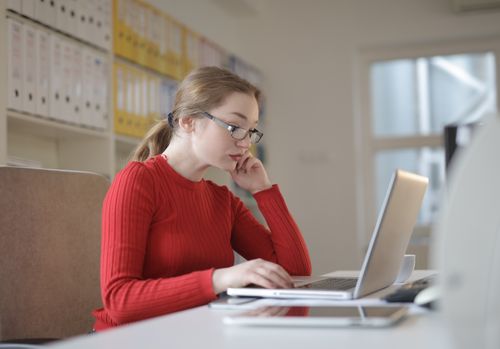 Free Focused woman in red sweater working on laptop at office desk with shelves Stock Photo