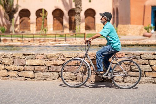 Free An elderly man rides a bicycle along a stone wall near historic Moroccan architecture. Stock Photo