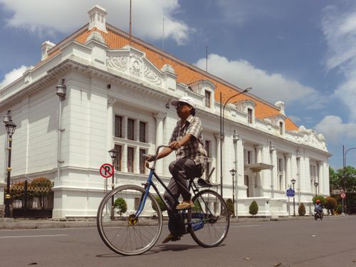 Free A man riding a bicycle past a historic colonial building in Surabaya, Indonesia, under a sunny sky. Stock Photo