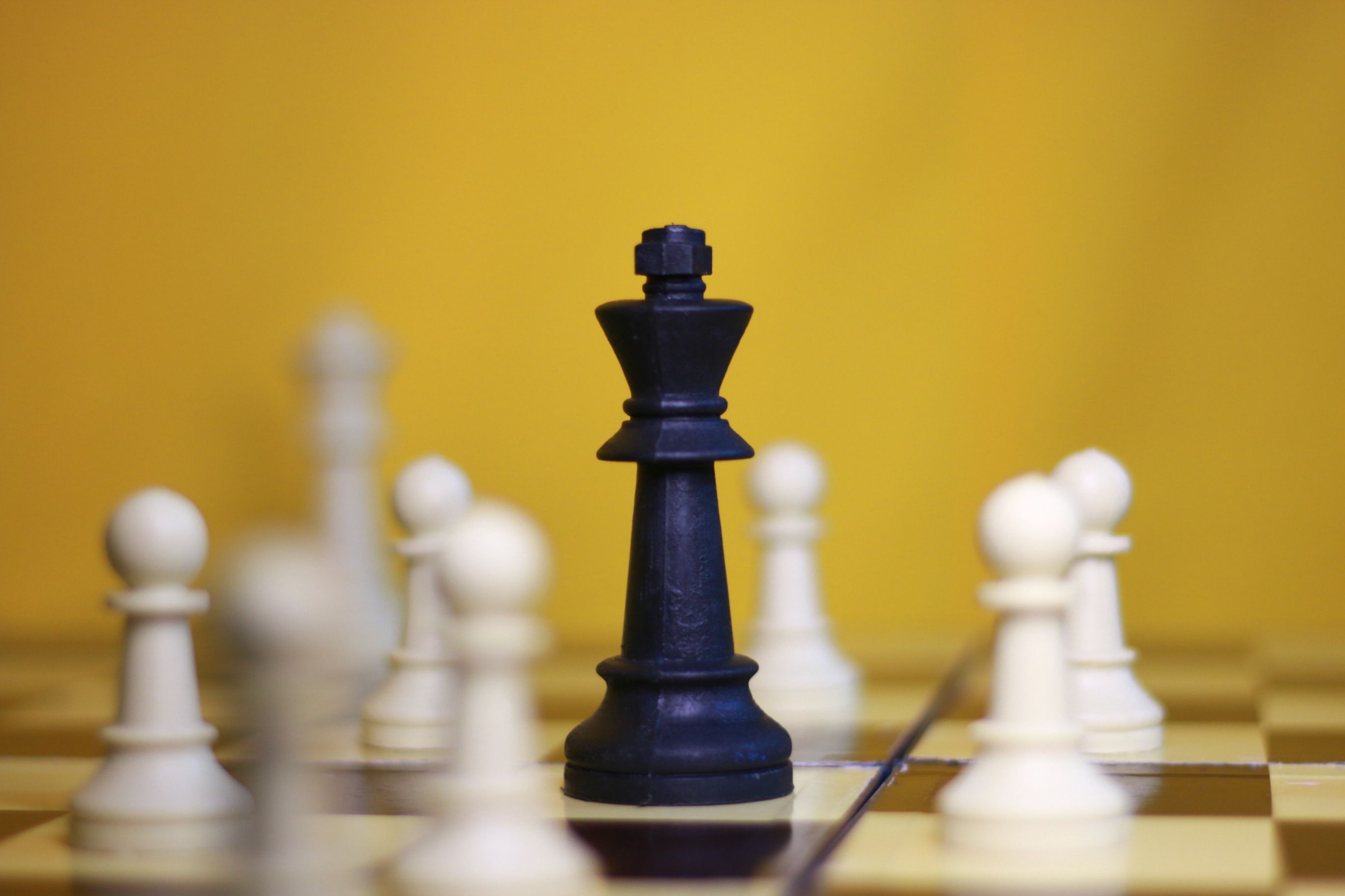 A black king chess piece surrounded by white pawns on a chessboard against a yellow background.