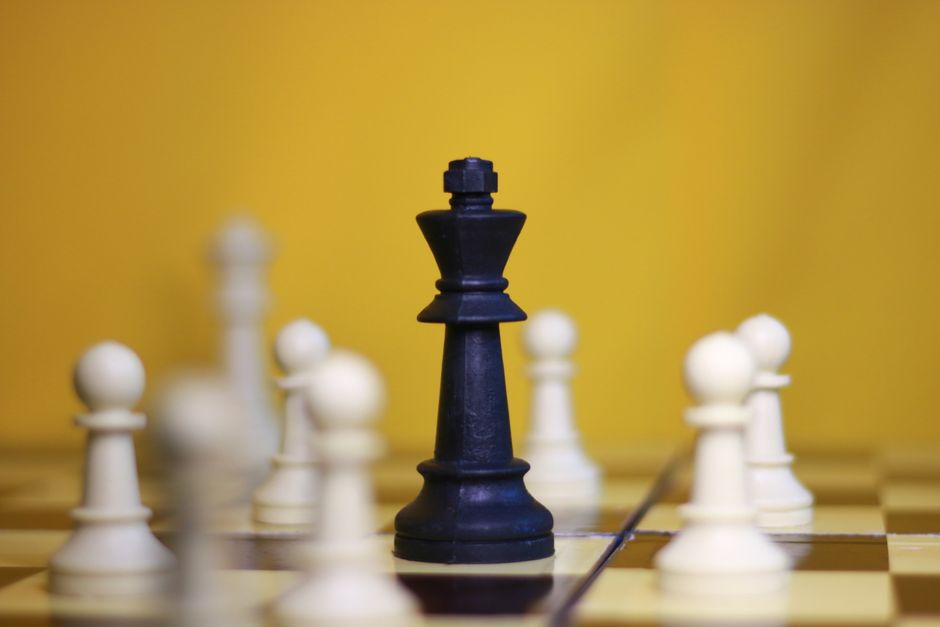 A black king chess piece surrounded by white pawns on a chessboard against a yellow background.