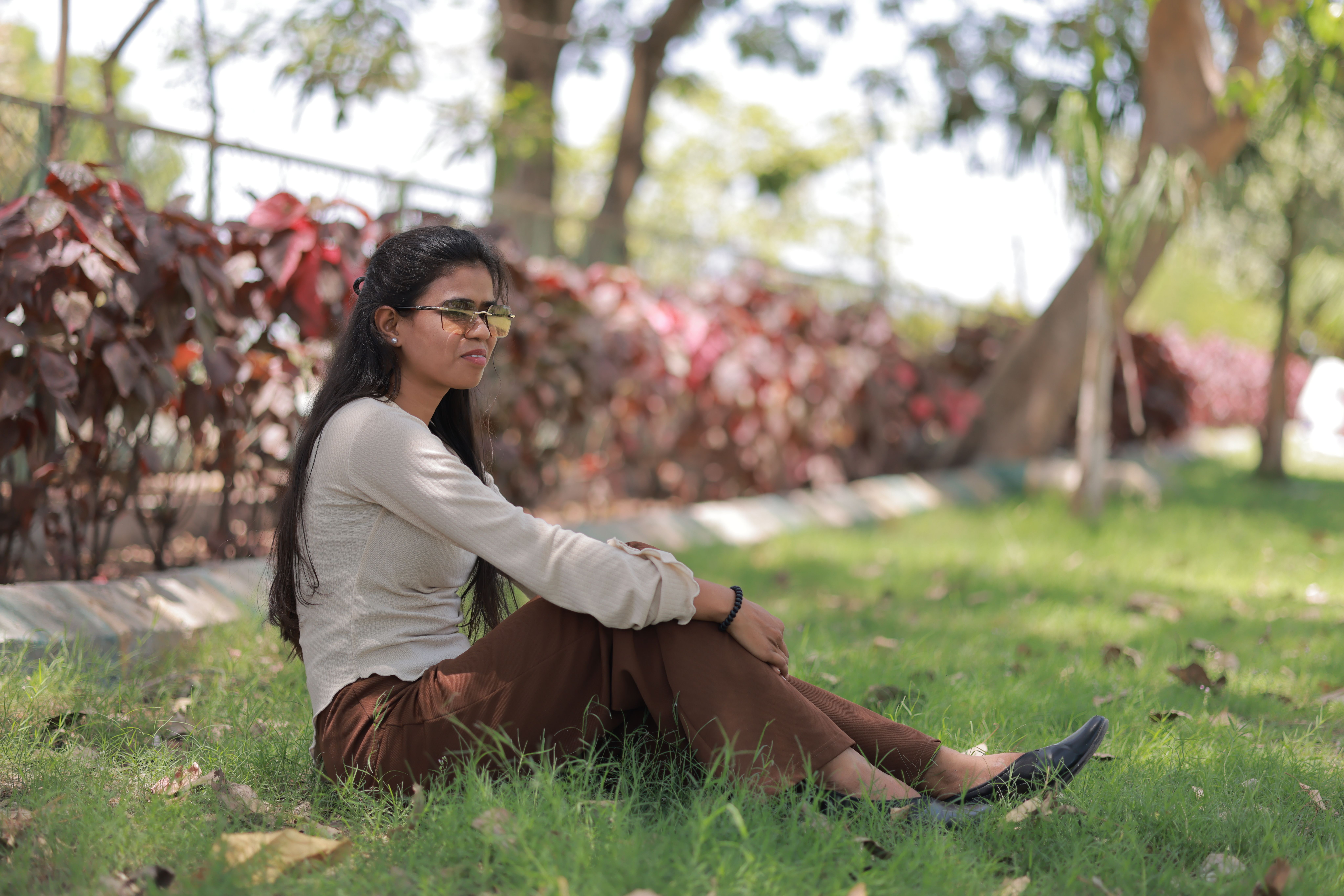 Young woman sitting on grass in a sunny park, surrounded by greenery and red leaves.