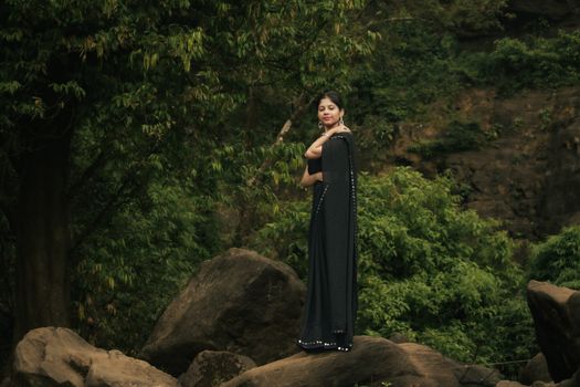 A woman in a black saree poses gracefully on rocks surrounded by lush greenery in a forest.