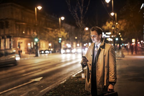 Foto Di Uomo In Cappotto Marrone Che Porta Una Valigetta E In Piedi Sul Marciapiede Di Notte Mentre Usa Il Suo Telefono