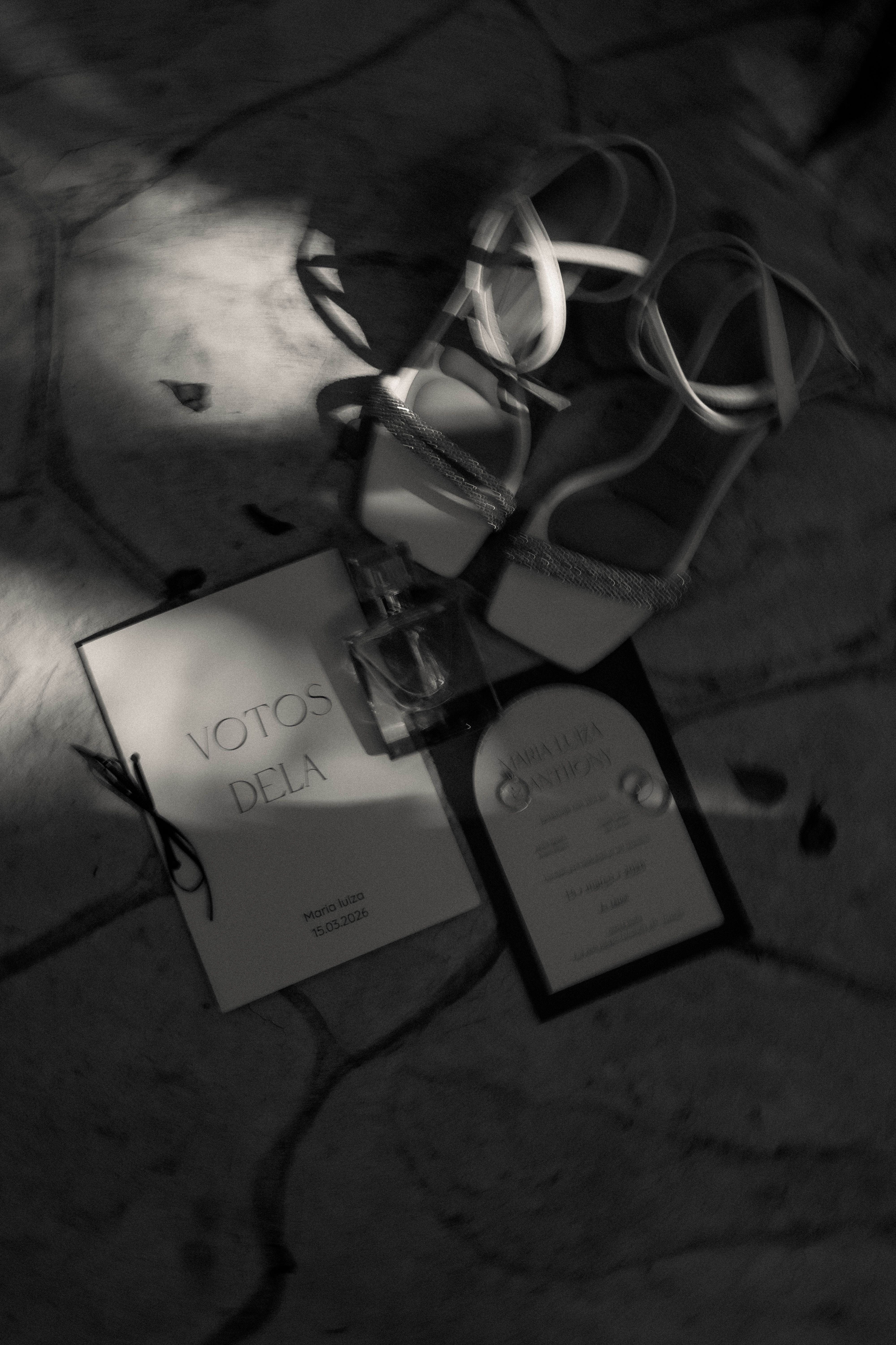 A monochrome flat lay of bridal essentials including vows book, invitation, perfume, and heels.