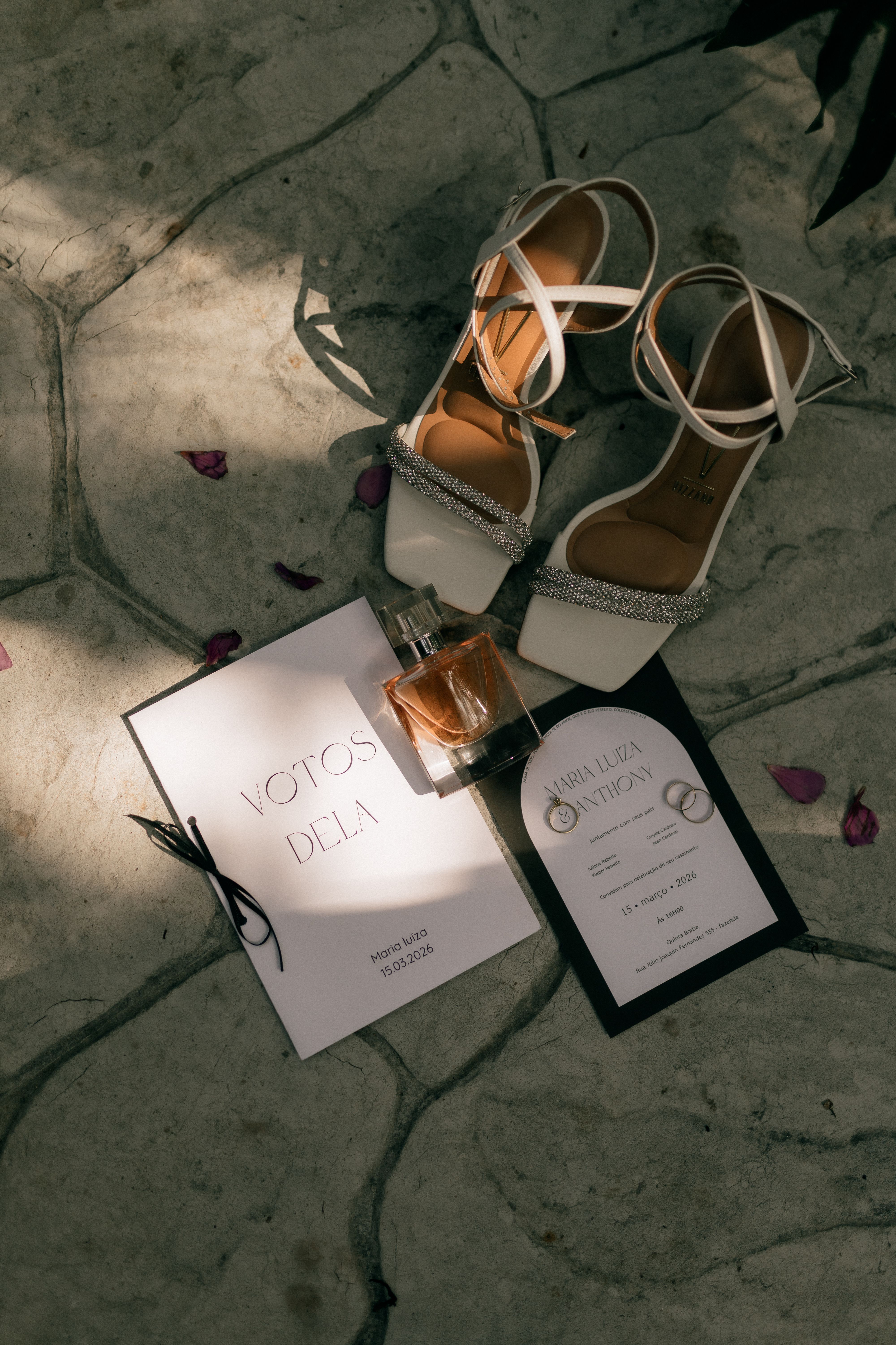 A wedding setup featuring shoes, vows book, invitation, and perfume under natural light.