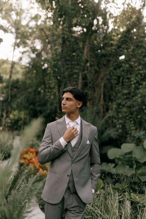 Free A young man in a grey suit poses in a lush outdoor garden setting, exuding elegance and style. Stock Photo