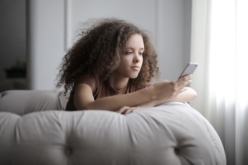Free A young woman with curly hair comfortably using her smartphone while sitting indoors. Stock Photo