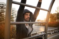 Young athletic male in sportswear with earphones doing exercises on ladder while training on sport ground near river in autumn morning in city