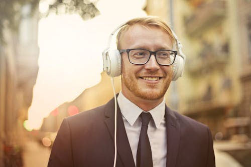 Free Selective Focus Photo of Smiling Man in a Black Suit and Black Framed Eyeglasses Listening to Music on White Headphones Stock Photo